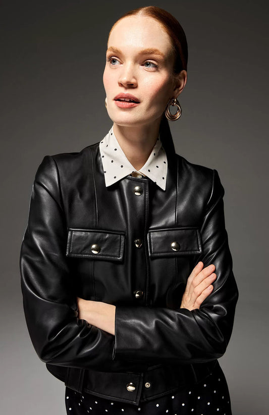 A woman with red hair tied back wears a black leather jacket over a white collared shirt with black polka dots. She has her arms crossed and looks to the side with a slight smile, standing against a dark background.