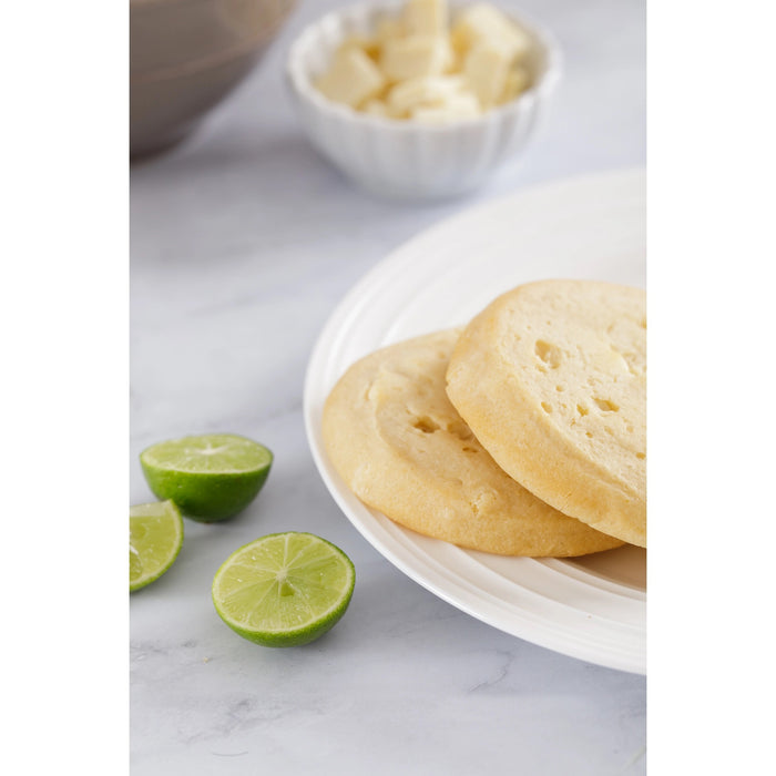 A package of WOW BAKING COMPANY Gluten Free Key Lime White Chocolate Cookie, in green and white, highlights its gluten-free, GMO-free status and natural ingredients, with tart lime and creamy white chocolate visible through the packaging.